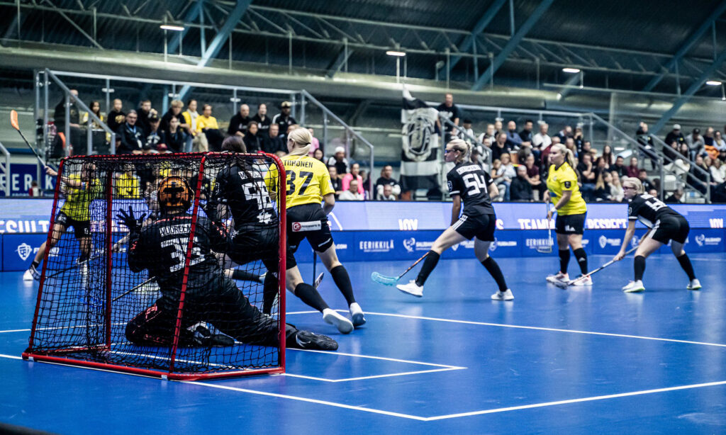 Kuva naisten salibandypelistä. Toisella joukkueella keltaiset paidat, toisella mustat. Kuvakulma toisen maalin takaviistosta. Kentällä näkyy useita pelaajia.