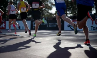 Kestävyysjuoksijoita radalla kaarteessa juoksemassa vastavaloon kuvattuna. Kuva rajattu niin, että juoksijoista osasta näkyy vain jalat lantiosta alaspäin, osasta koko vartalo. Juoksijoiden varjoja heijastuu maahan.