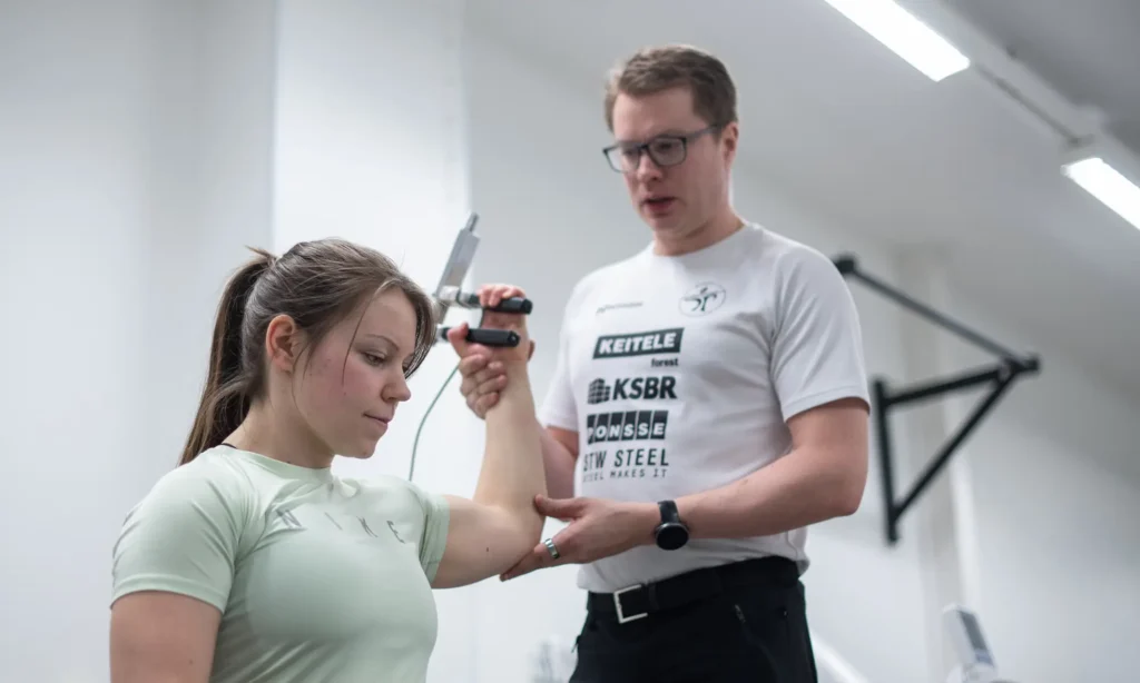 Fysioterapeutti tekee naiskeihäänheittäjälle puristusvoimatestiä. Heittäjä istuu vasen käsi hartiantasolla kyynärpää 90 asteen kulmassa kädessään sormien puristusvoimaa mittaava laite. Fysioterapeutti (mies) pitää kiinni heittäjän kädestä ja mittarista.