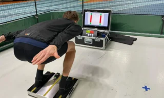 Mäkihyppääjä kyykyssä laskuasennossa voimalevyantureiden päällä. Edessä lattialla hänelle on mittausjärjestelmä ja näyttö, jolta hän seuraa jalkojen voimajakaumaa.