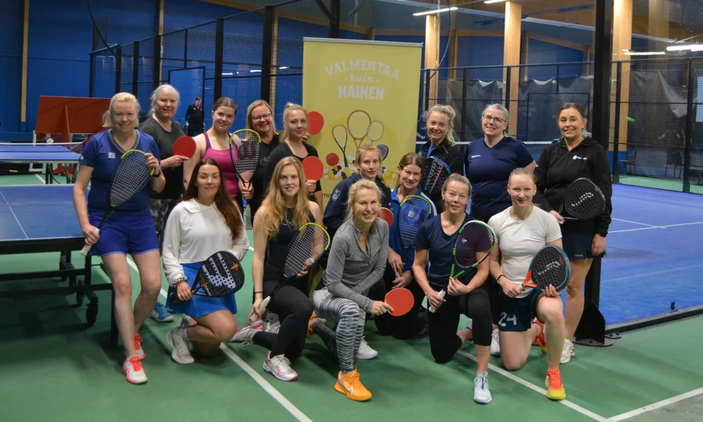 Joukko urheilullisen näköisiä naisia yhteiskuvassa. Jokaisella kädessään jokin pallopelimaila. Taustalla näkyy padelpelikenttiä ja pöytätennispöytiä.
