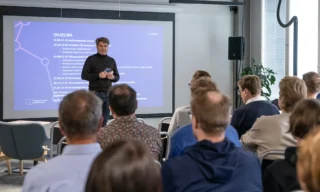 Kuva suomalaisen urheilun TKI-verkostopäivästä Jyväskylässä 17.4.2024. Kuvassa KIHUn johtaja Arto Kuusisto puhuu osallistujille. Hänen taustalla näkyy päivän ohjelma seinälle heijastettuna.