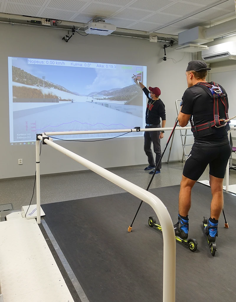 Hiihtäjä seisoo juoksumatolla ja katsoo seinälle, jonne on heijastettu kuvaa todellisesta kilpailureitistä. Kuva niin sanotusta simulaatioympäristöstä Vuokatista.