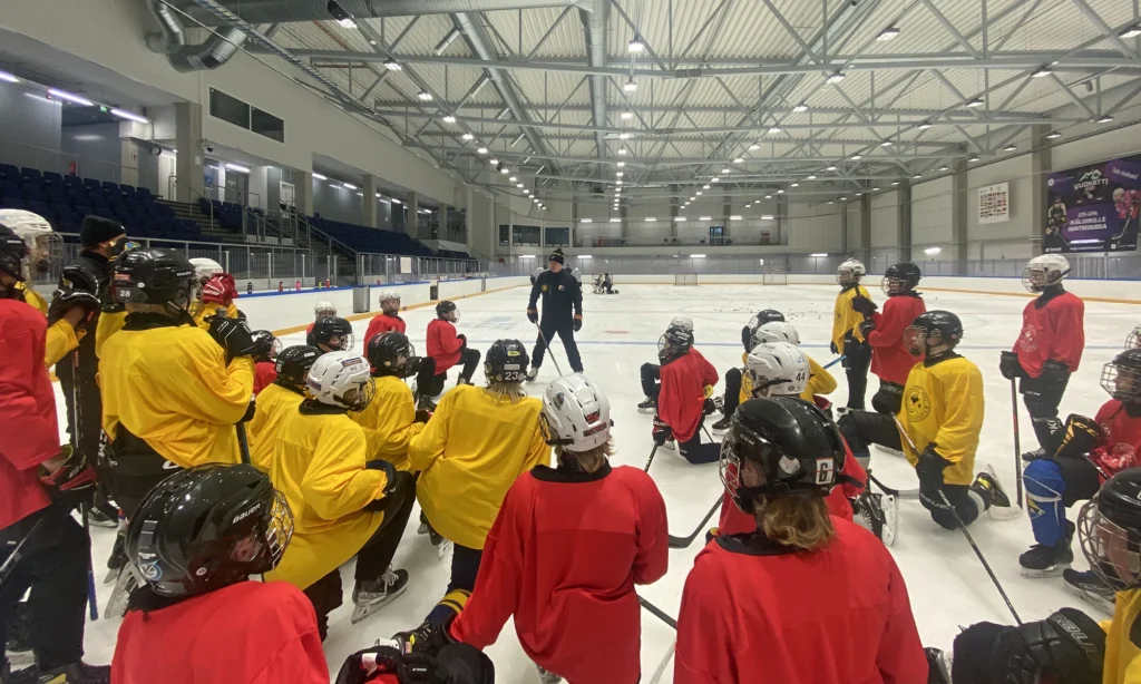 Joukko jääkiekkoilijoita punaiset tai keltaiset treenipaidat päällään polviasennossa jäällä kuuntelemassa valmentajan puhetta. Taustalla näkyy muuten tyhjä jääkiekon harjoitushalli.