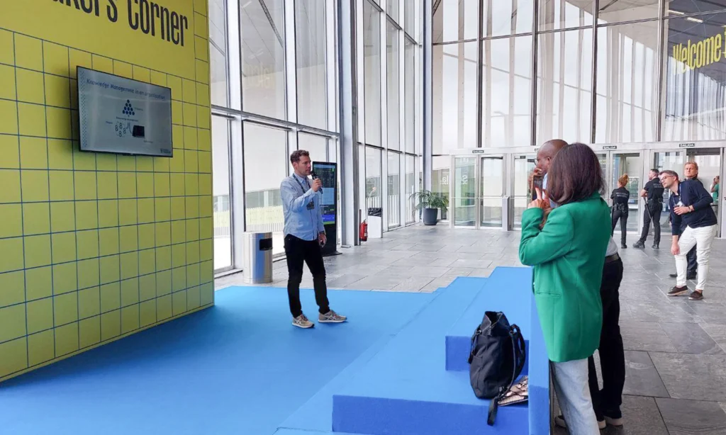 Mieshenkilö mustissa farkuissa ja vaalean sinisessä kaulupaidassa pitämässä seminaariesitystä avoimessa hallitilassa. Taustalla paljon isoja ikkunoita, esiintymispaikan keltainen tausta. Lattia on esiintymislavan kohdalla sininen. Yleisöä näkyy vain vähän kuvan reunassa.