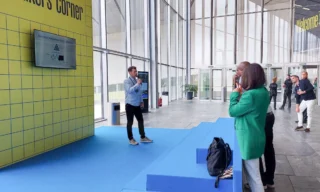 Mieshenkilö mustissa farkuissa ja vaalean sinisessä kaulupaidassa pitämässä seminaariesitystä avoimessa hallitilassa. Taustalla paljon isoja ikkunoita, esiintymispaikan keltainen tausta. Lattia on esiintymislavan kohdalla sininen. Yleisöä näkyy vain vähän kuvan reunassa.