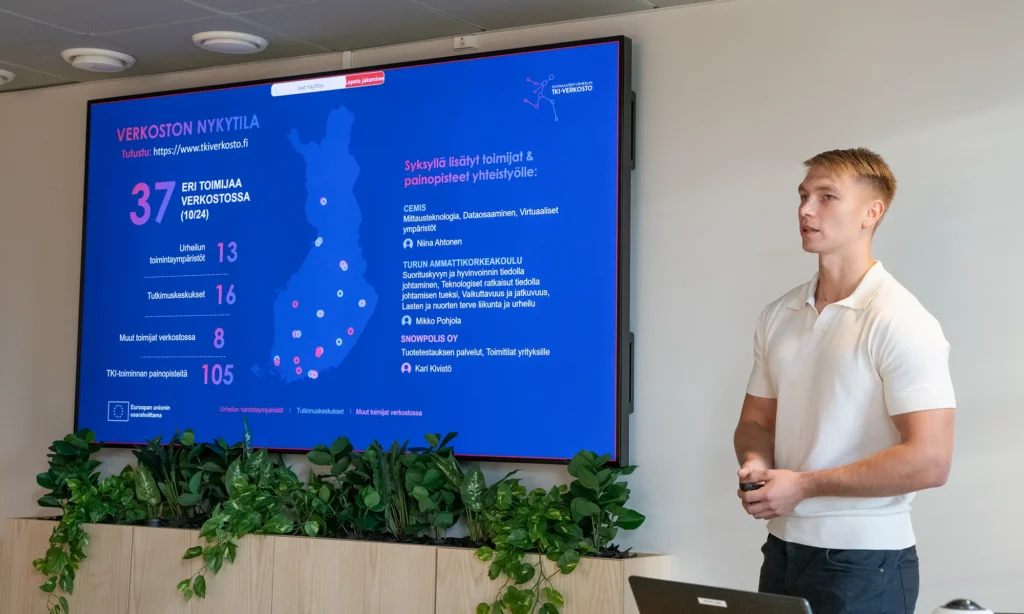 Mieshenkilö pitämässä esitystä seminaariyleisölle. Valkoinen paita päällään. Taustalla iso näyttö, jossa sinisävyinen karttakuva urheilun tki-toimijoista Suomessa.