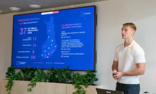 Mieshenkilö pitämässä esitystä seminaariyleisölle. Valkoinen paita päällään. Taustalla iso näyttö, jossa sinisävyinen karttakuva urheilun tki-toimijoista Suomessa.