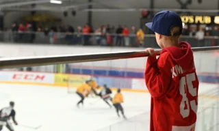 Poika, jolla on punainen pelipaita päällään ja sininen lippis päässään, katsoo jääkiekkopeliä jäähallissa nojaten kaiteeseen.