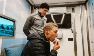 Hiihtäjä on tekemässä puhallustestiä hiihtomaajoukkueen rekassa yskätutkimukseen liittyen. Tutkija Mäki-Heikkilä seuraa taustalla.