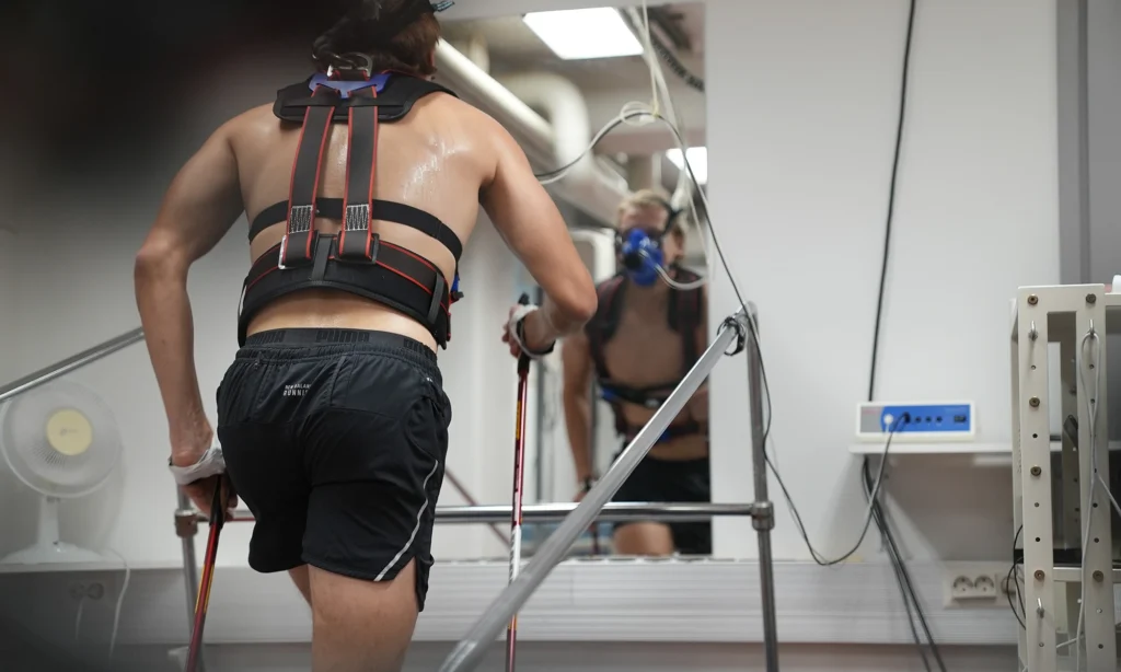 Urheilija sauvakävelytestissä juoksumatolla. Kuvattuna takaapäin. Mieshenkilö, mustat sortsit, ei paitaa.