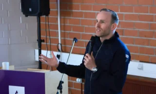 Mika Poutala puhumassa seminaariyleisölle salin edessä. Toisessa kädessä mikrofoni. Taustalla tiiliseinää.