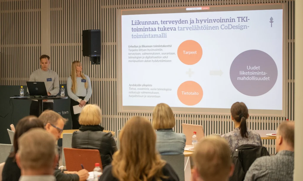 Mies ja nainen pitämässä esitystä yleisön edessä. Seinälle on heijastettuna CoDesingSPORT-hankkeen infoa.