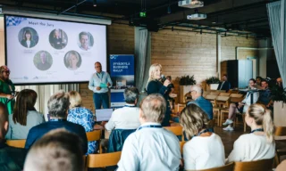 Nainen puhuu mikrofoniin yleisön edessä. Taustalla seinälle heijastettuna esiintymiseen liittyvää materiaalia.